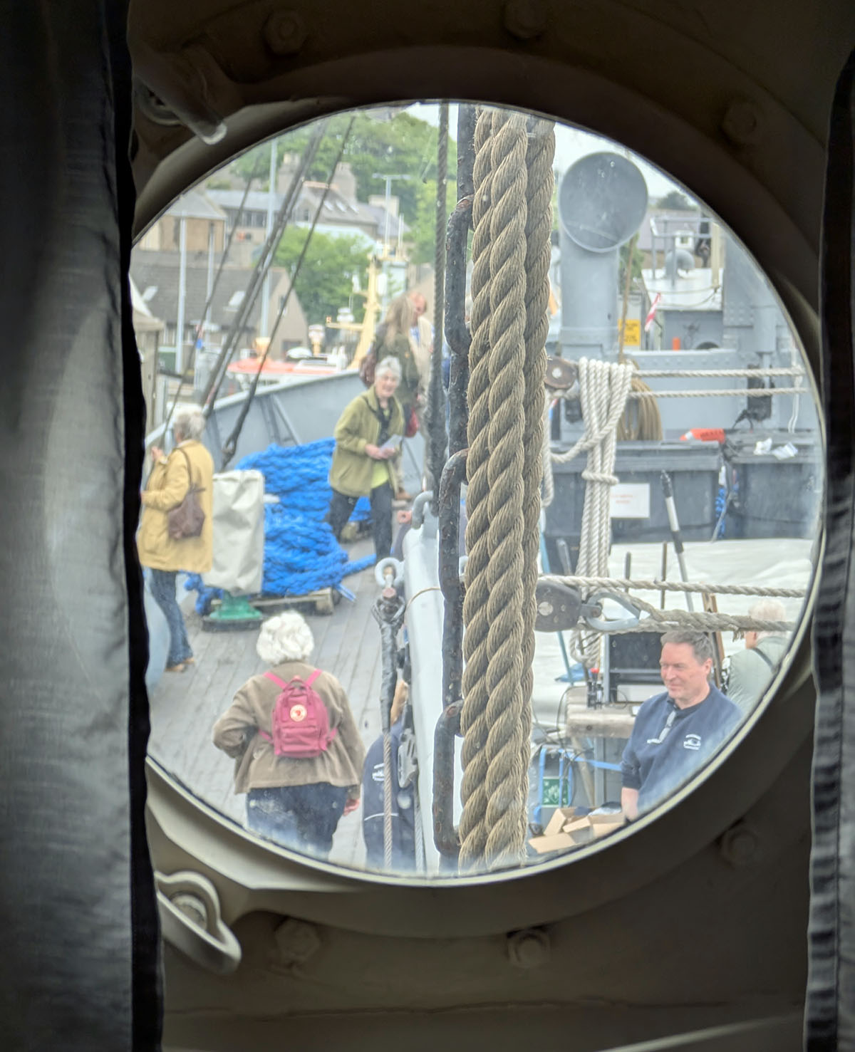 View through a porthole showing volunteers and visitors on the outside deck. The ship is painted wartime grey.